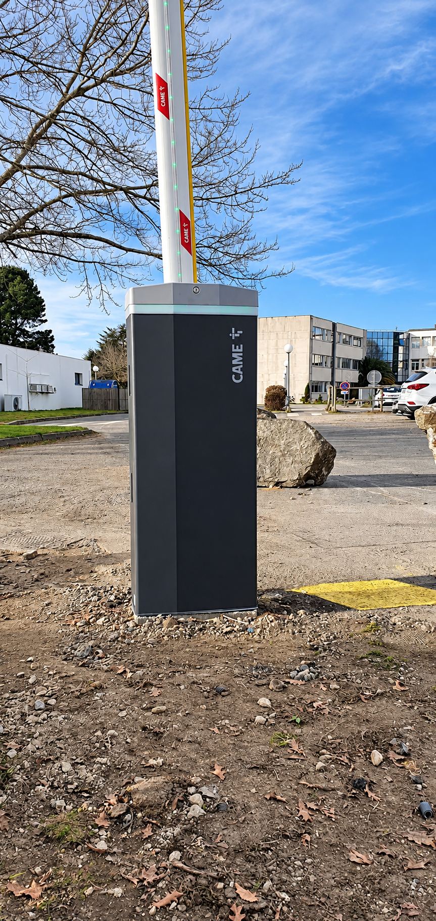 Une barrière levante automatique de marque CAME, avec sa lisse blanche relevée, est installée sur un parking en terre devant des bâtiments de bureaux.