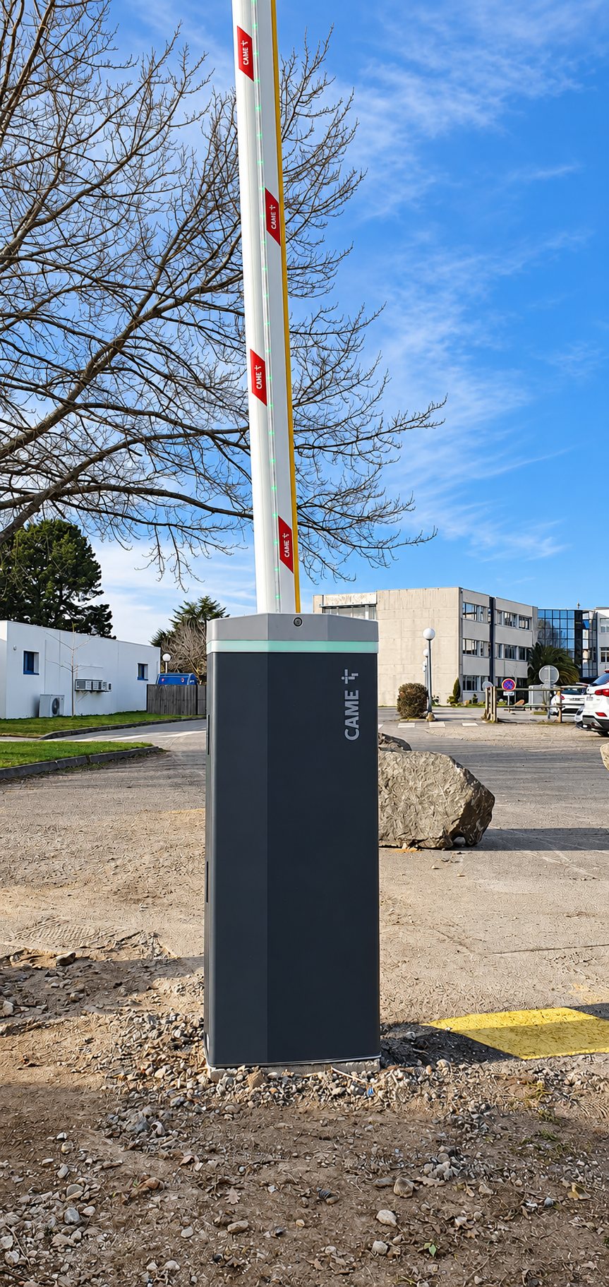 Une barrière automatique CAME à la lisse relevée, posée sur un parking en terre devant un rocher et des bâtiments, sous un ciel bleu.