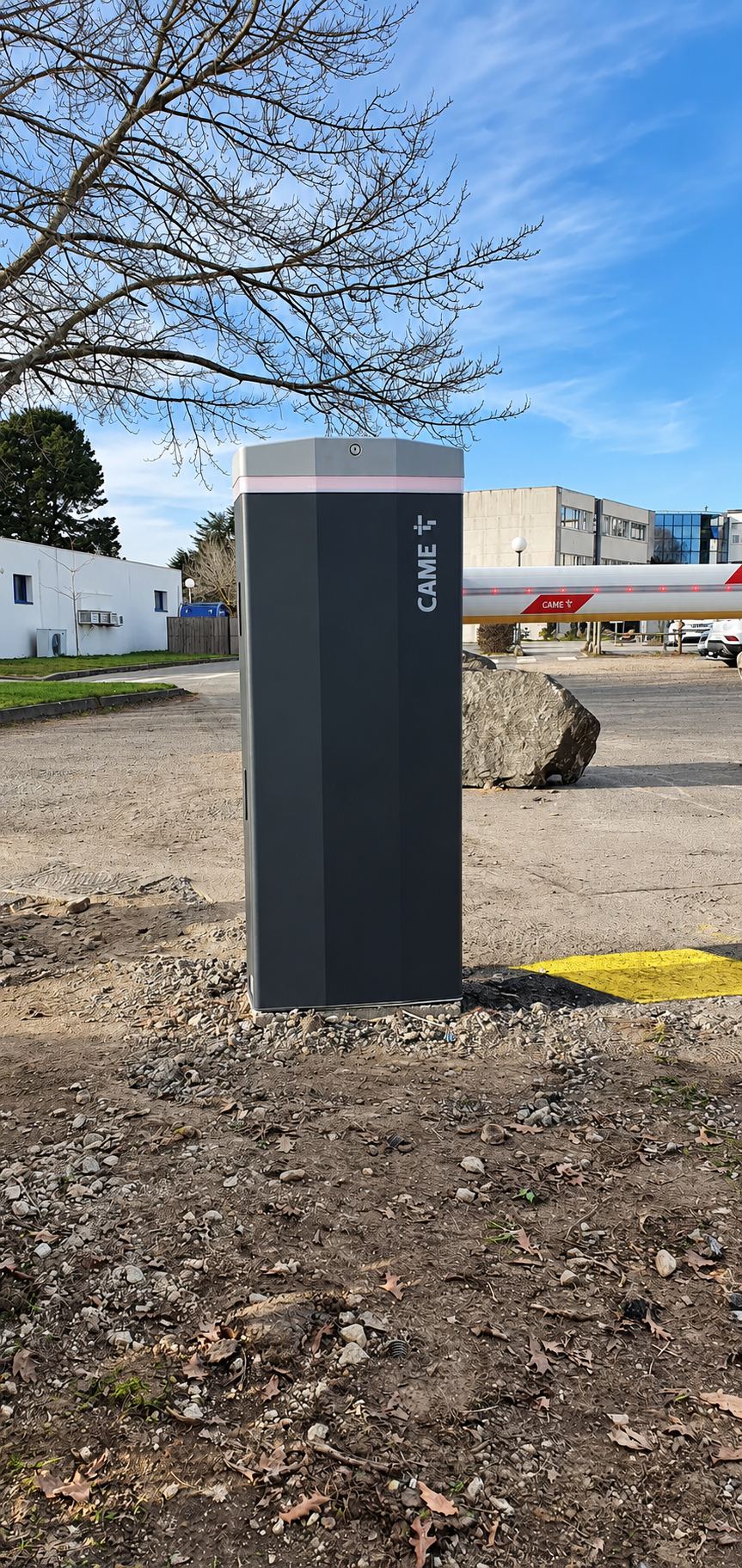 Une borne grise de barrière automatique de marque CAME se dresse sur un parking en terre, avec une lisse rouge et blanche à l'arrière-plan.
