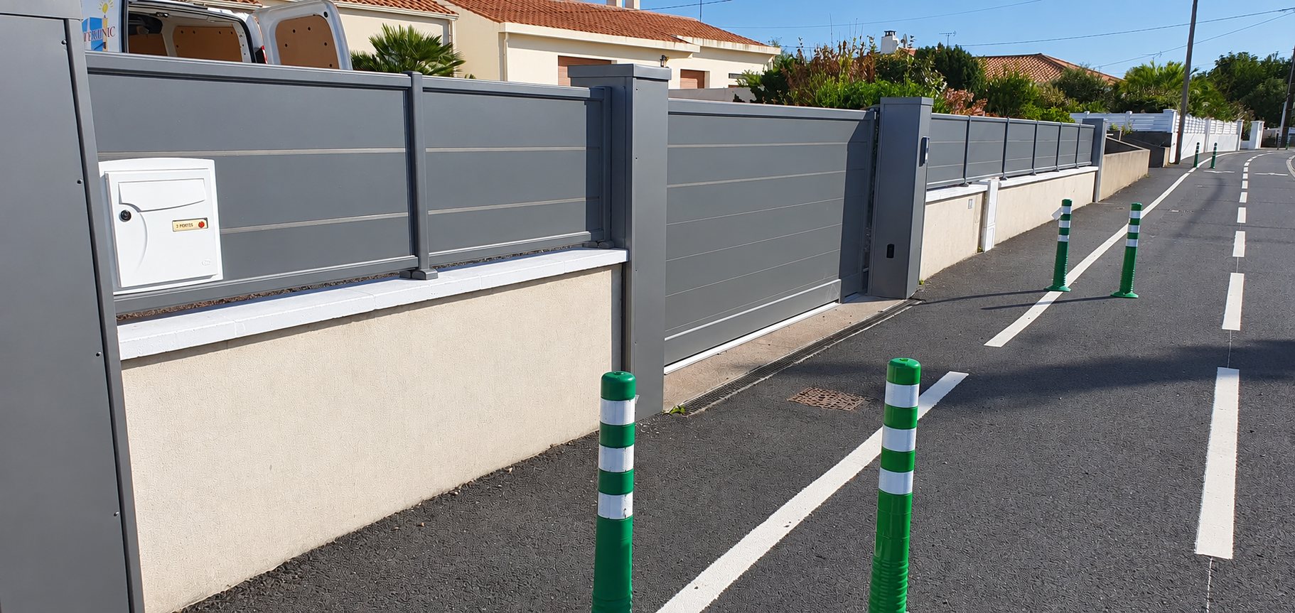 Une clôture et un portail coulissant en aluminium gris sur un muret blanc bordent une rue avec des balises vertes et blanches au premier plan.