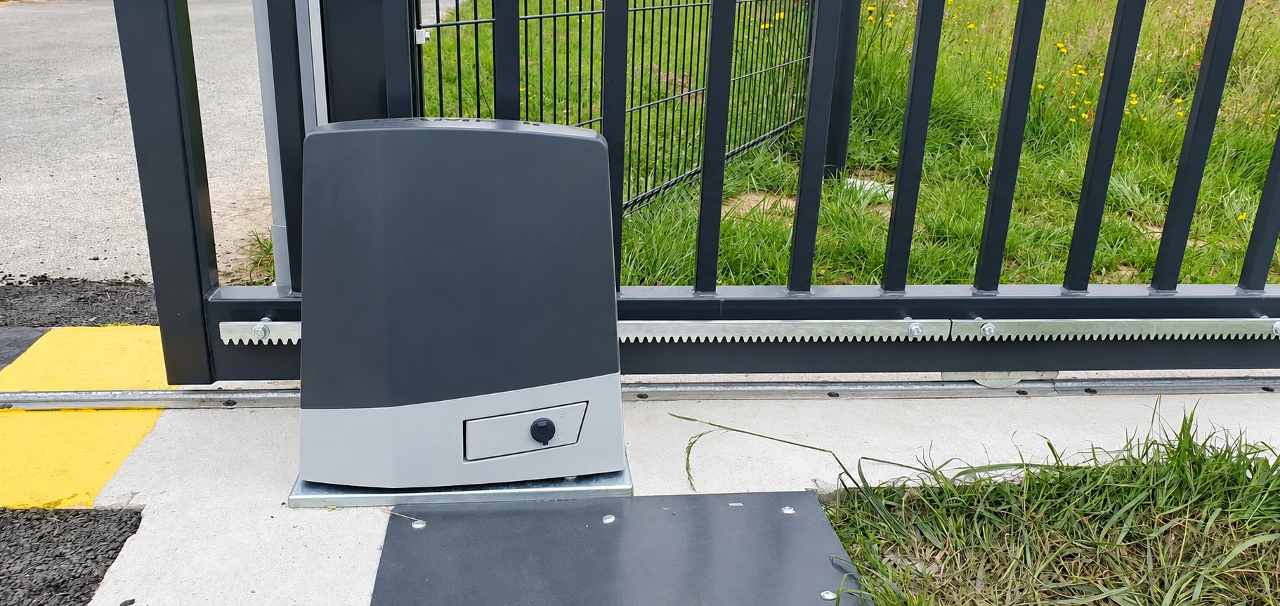 Un moteur gris et noir pour portail coulissant est fixé sur une platine au sol, à côté de la crémaillère d'un portail noir et d'une pelouse.