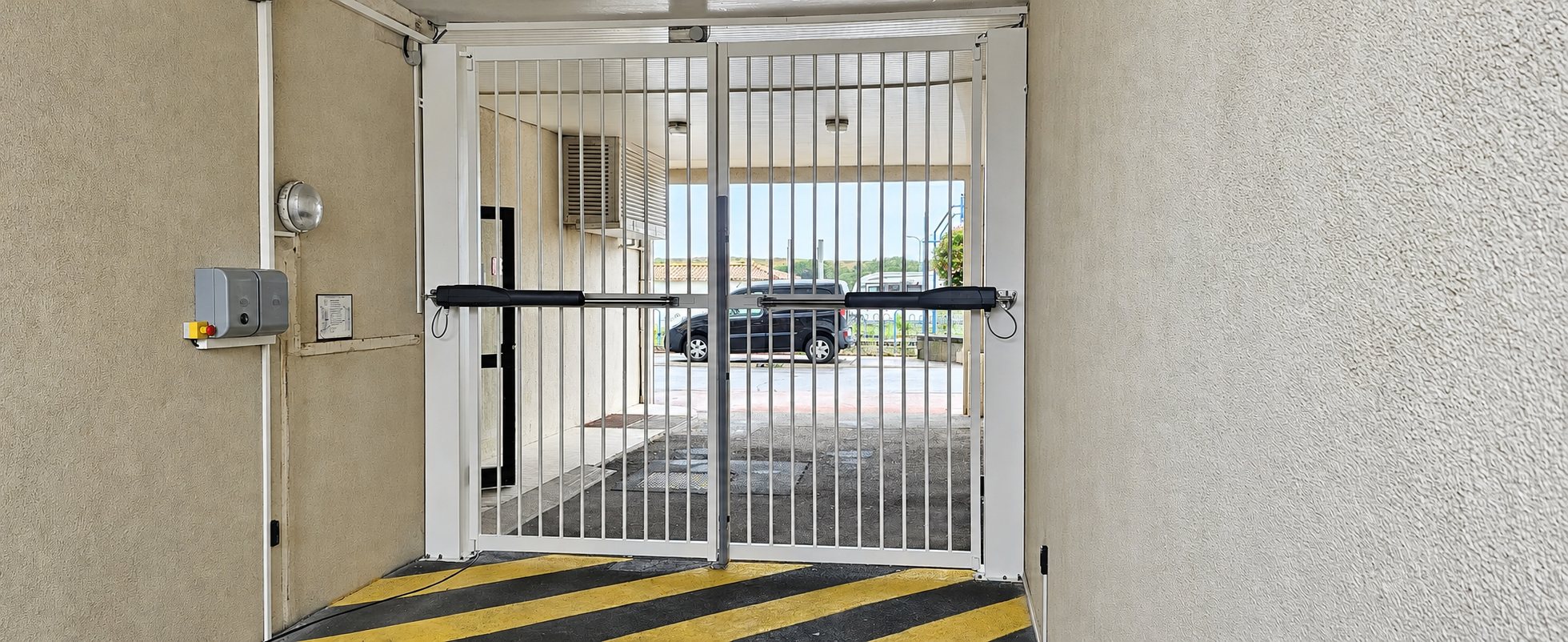 Un portail à barreaux blanc à deux battants, équipé de vérins automatiques, ferme un passage couvert donnant sur un parking où stationne une voiture.