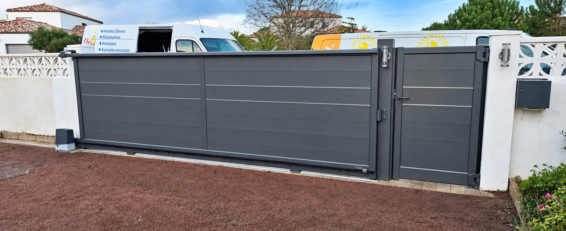 Un portail coulissant gris avec son portillon assorti, posé entre deux murs blancs, avec des camionnettes d'entreprise garées derrière.