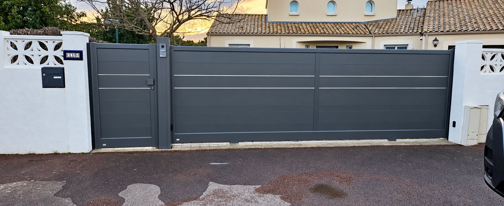 Un portail coulissant et un portillon en aluminium gris anthracite, entre deux murets blancs décoratifs, devant une maison à toit de tuiles.