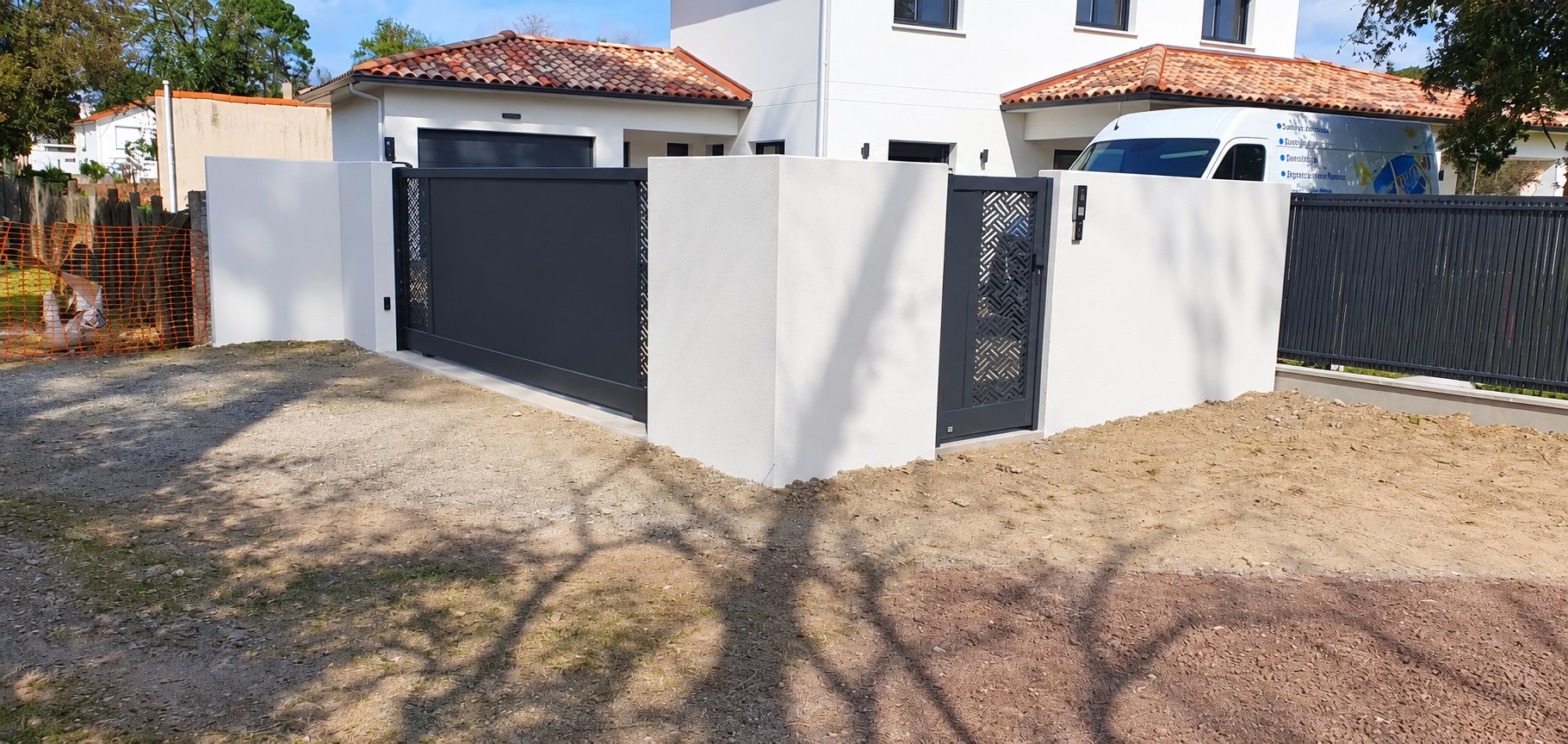 Un portail coulissant et un portillon ajouré gris anthracite encadrés de murets blancs devant une maison moderne avec une camionnette garée derrière.