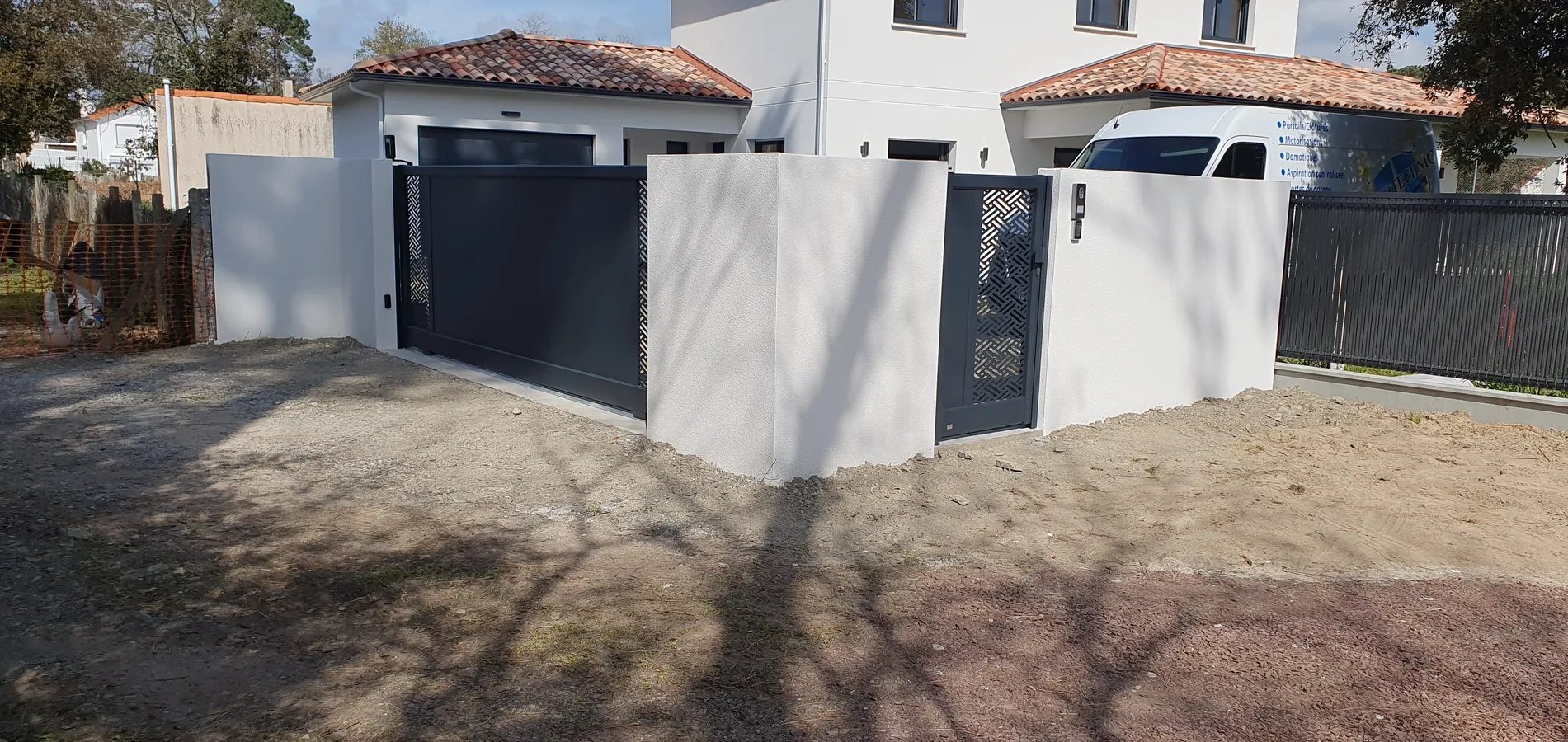 Un portail coulissant et un portillon en aluminium gris anthracite à motifs ajourés, encadrés de murets blancs, devant une maison moderne avec une camionnette garée à droite.