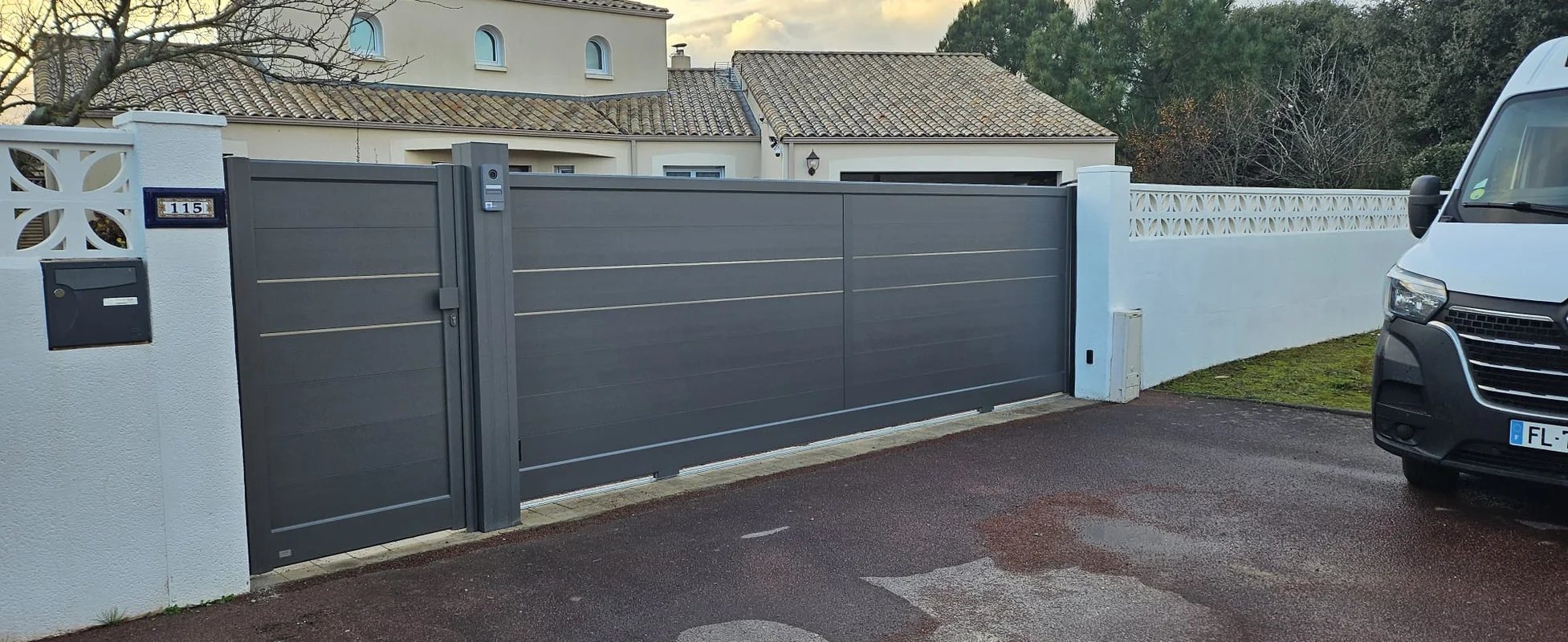 Un portail coulissant et un portillon en aluminium gris anthracite installés entre des murets blancs devant une maison, avec une camionnette blanche garée à droite.