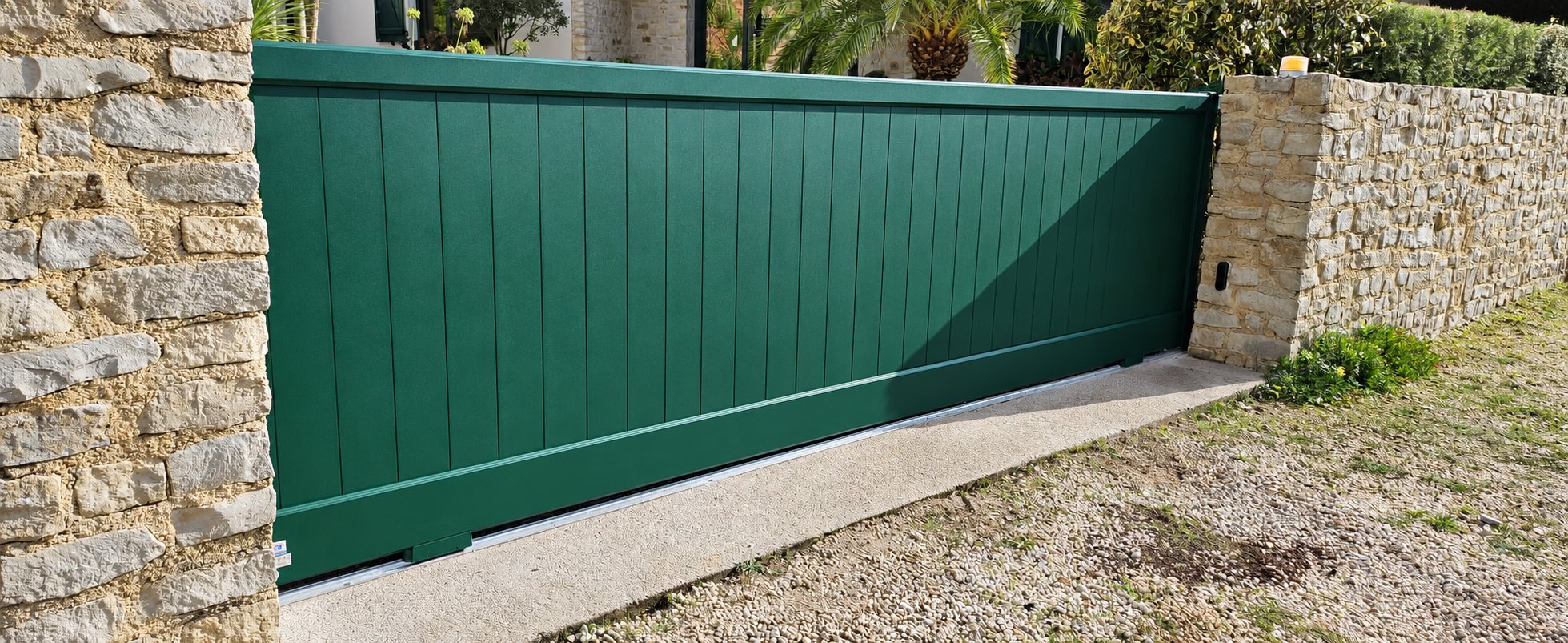 Un portail coulissant en aluminium vert à lames verticales, encadré par des murets en pierre naturelle, avec un voyant orange au sommet du pilier.