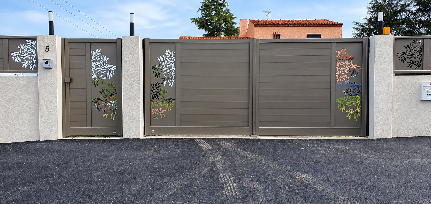 Un portail coulissant et un portillon en aluminium gris taupe ornés de motifs de feuilles découpées, entre des piliers blancs devant une maison à toit de tuiles.