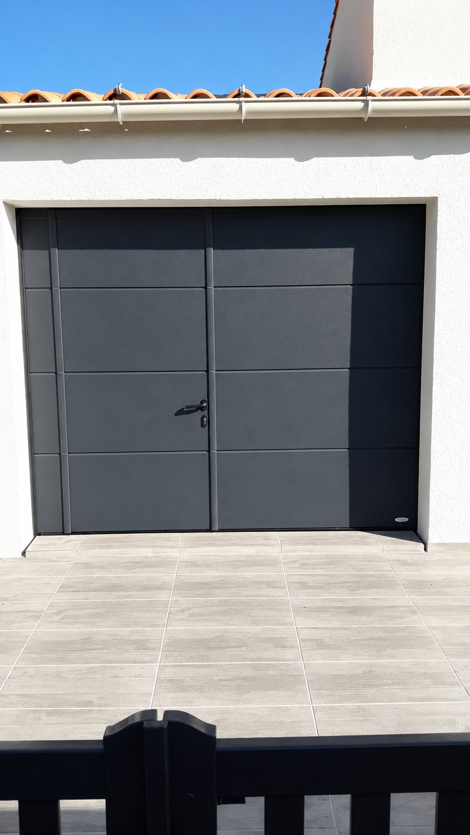 Porte de garage sectionnelle gris anthracite avec portillon intégré, installée sur une façade blanche au-dessus d'une terrasse carrelée et derrière un portail noir.