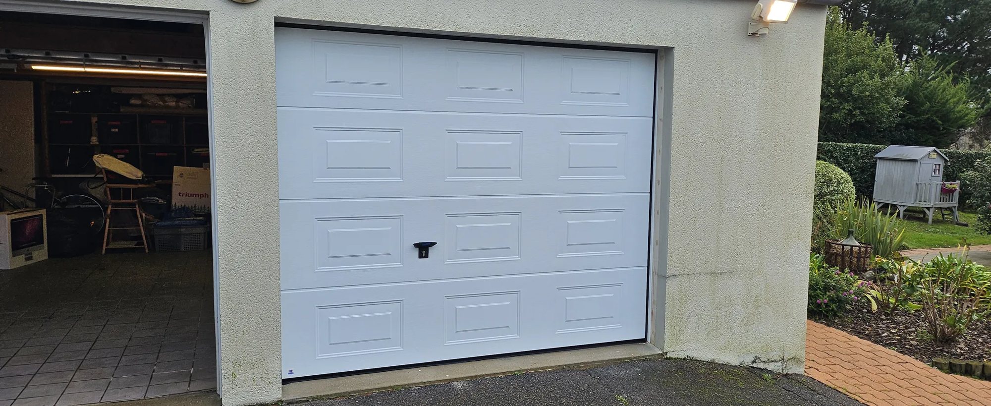 Une porte de garage sectionnelle blanche à panneaux est installée dans la façade crépie d'une maison, à côté d'un second garage ouvert et d'un jardin avec une cabane.