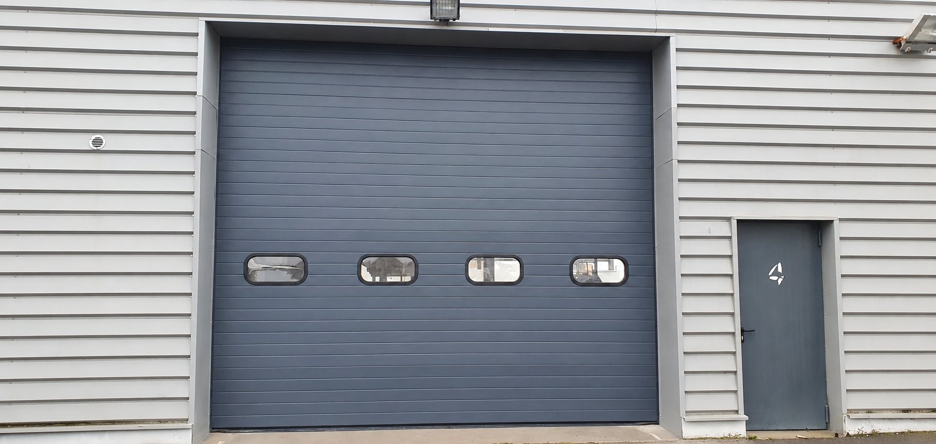 Grande porte sectionnelle grise munie de quatre petits hublots, à côté d'une porte piétonne grise numérotée 4, sur une façade au bardage gris clair.