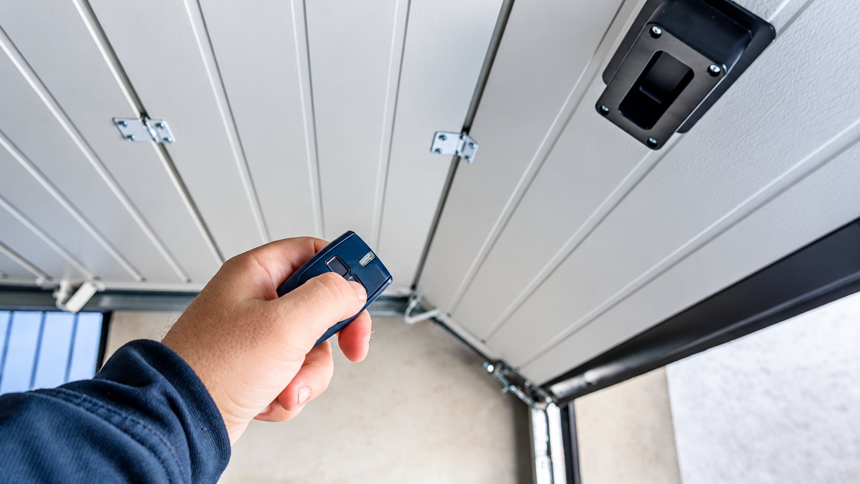 Une main tient une petite télécommande bleue pour ouvrir une porte de garage sectionnelle blanche vue de l'intérieur.